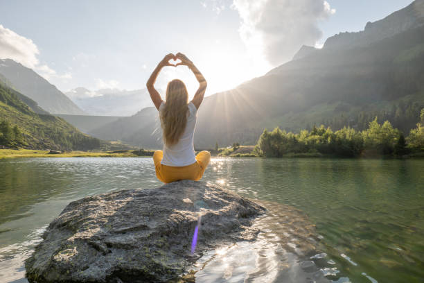 une jeune femme est assise sur un rocher au-dessus du lac et fait un cœur avec les mains - mountain rock sun european alps photos et images de collection