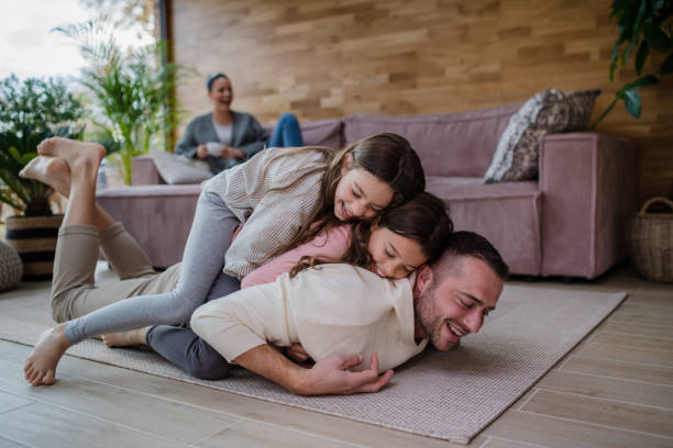 zwei glückliche schwestern, die spaß mit dem vater haben, auf dem rücken liegen, mit der mutter im hintergrund zu hause - couple loving lying on back carpet stock-fotos und bilder