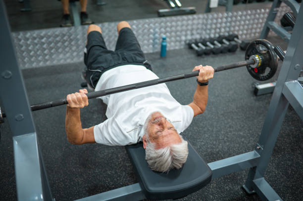 Senior Caucasian man in late 60s doing a bench press exercise while having a workout in the gym Senior Caucasian man in late 60s lifting weights by doing a bench press exercise in the gym to stay fit and healthy during his retirement senior bodybuilders stock pictures, royalty-free photos & images