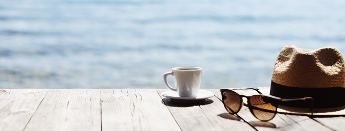Coffee cup, sunglasses and straw hat on a table at mediterranean cafe. Travel, tourism, vacations, holidays concept