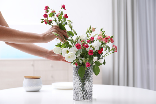 Woman taking beautiful flowers from vase on white table in room, closeup