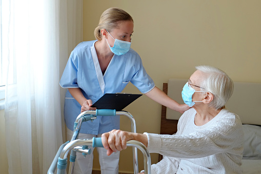 Hospital nurse wearing a face mask, holding a clipboard with medical history for an elderly lady. Senior woman and her designated care giver discussing test results. Background, close up, copy space.