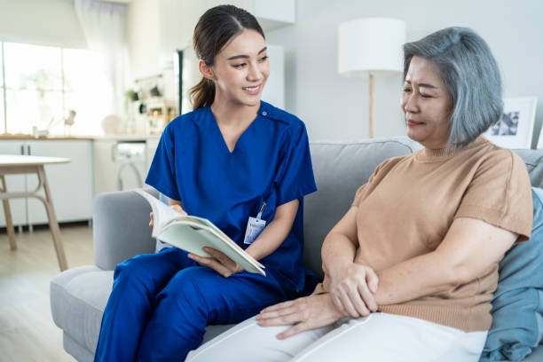 Asian woman nurse taking care of senior elderly female at nursing home. Young Caregiver Therapist doctor sit on sofa, read a book to happy older handicapped patient. Medical insurance service concept. stock photo