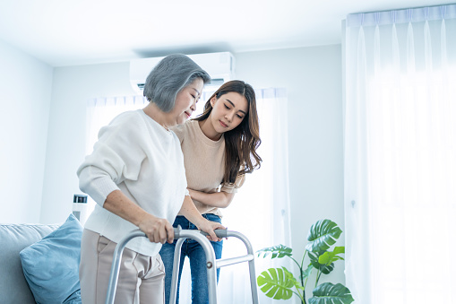 Asian daughter support older woman grandma walk with walker at home. Beautiful girl help and take care of senior elderly mature mother patient stand and doing physical therapy in living room in house.