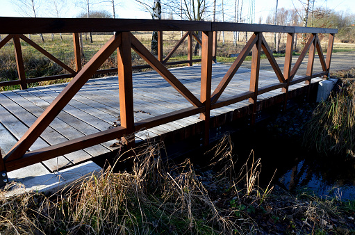 a park bridge with a wooden railing leads across the stream. The beams are fixed to a steel crossbeam that holds in a concrete foundation. road from gravel, in front of the bridge barrier, bolts and nuts, crossbeam, polder, river, pond