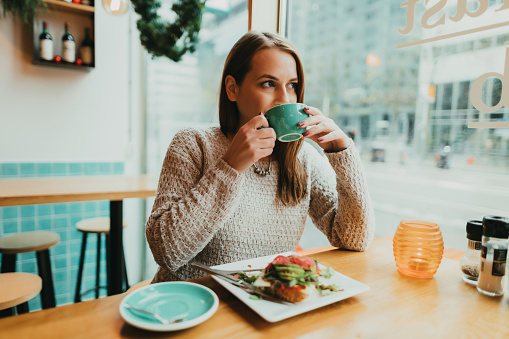 Young woman sitting in the coffee bar, drinking coffee from a blue mug and eating healthy vegan avocado toast for breakfast