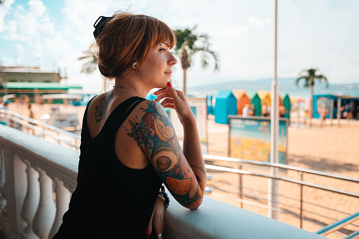 Portrait of a young smiling woman with a tattoo on her arm, looking at the sea. There is a beach in the background. The concept of summer vacation.