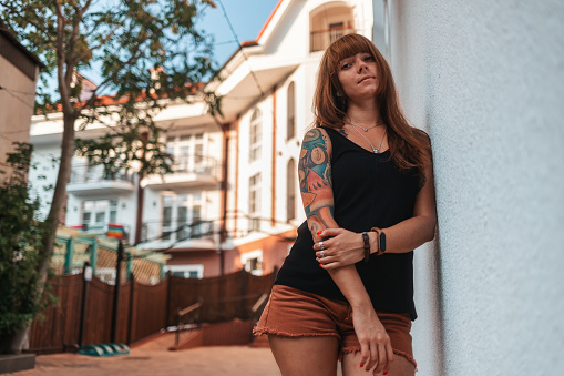 A pretty young woman with tattoos is leaning against the wall of the house. Bottom view. The concept of waiting and loneliness.
