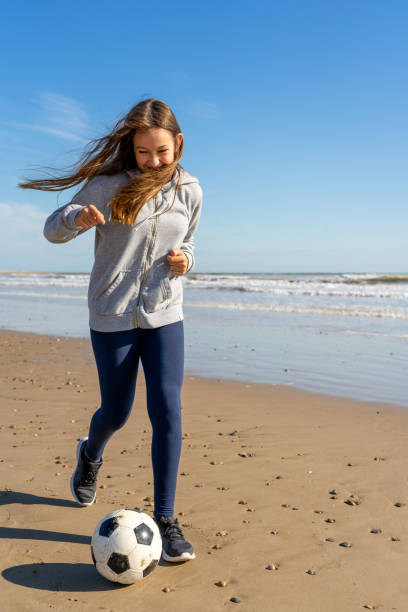 dorastająca hiszpanka biega po plaży z piłką nożną, podczas gdy jej włosy zakrywają twarz przed wiatrem. koncepcja ćwiczeń i zabawy. - hiszpanka_& zdjęcia i obrazy z banku zdjęć