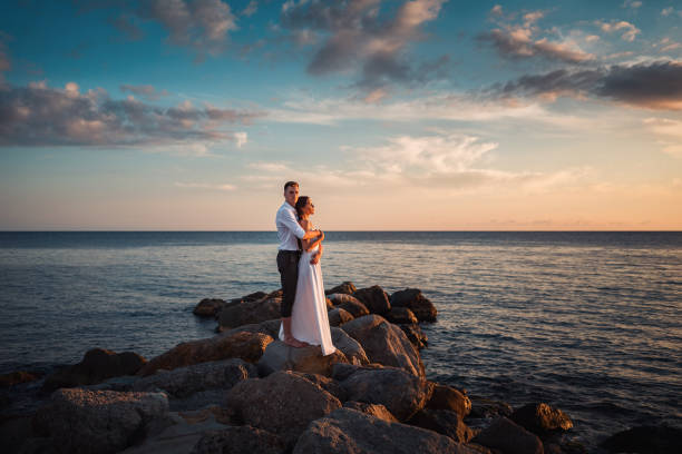 結婚式の日。新婚夫婦は海岸の岩の上に立って抱き合い、ポーズをとります。背景には、夕日と海と空が雲の中にある - weding gown ストックフォトと画像