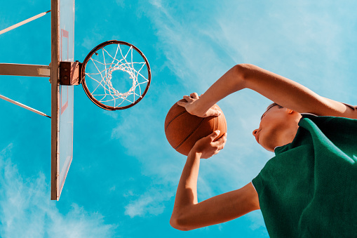 Deportes y baloncesto. Un joven adolescente en chándal verde lanza una pelota a la canasta. Vista desde abajo. Cielo azul al fondo photo