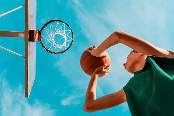 Photo of Sports and basketball. A young teenager in a green tracksuit throws a ball into the basket. Bottom view. Blue sky in the background