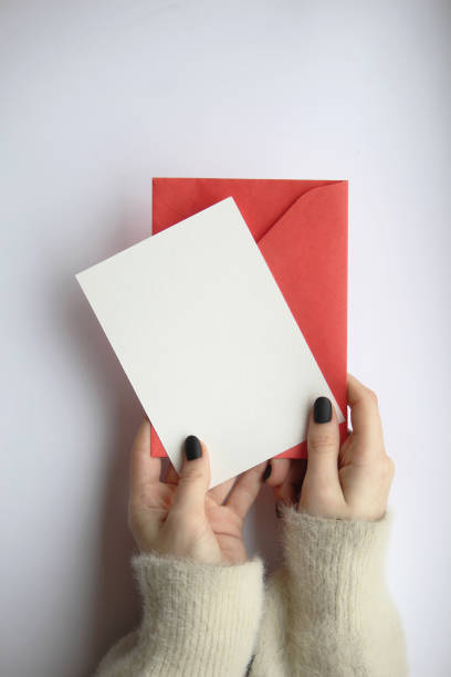 istock female hands in a sweater with manicure, holding a white sheet and a red envelope on a white background. postcard layout 1363811654