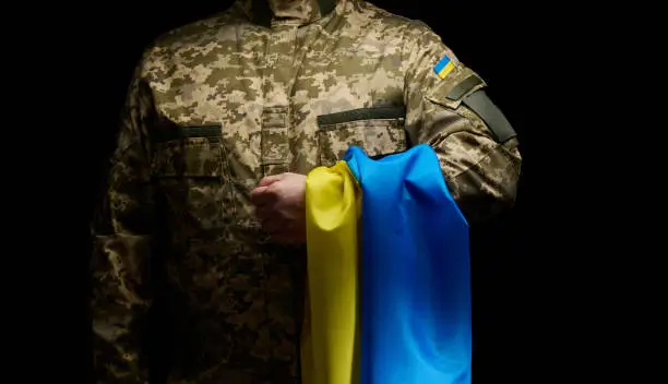 Photo of a soldier of the Ukrainian armed forces stands with a blue-yellow flag of Ukraine on a black background. Honoring veterans and commemorating those killed in the war