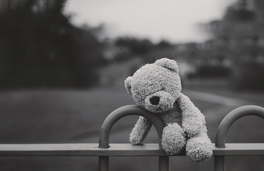 Oso de peluche perdido en blanco y negro sentado en el banco en el patio de recreo en un día sombrío, muñeca de oso de cara solitaria y triste acostada sola en el parque, juguete perdido, concepto de soledad, Día Internacional de los Niños Desaparecid photo