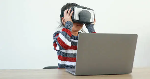 Photo of The little girl wearing VR sitting at desk in the home glasses virtual Global Internet connection metaverse. Future kids in white clothing wearing VR headsets for a game. concept advanced technology.