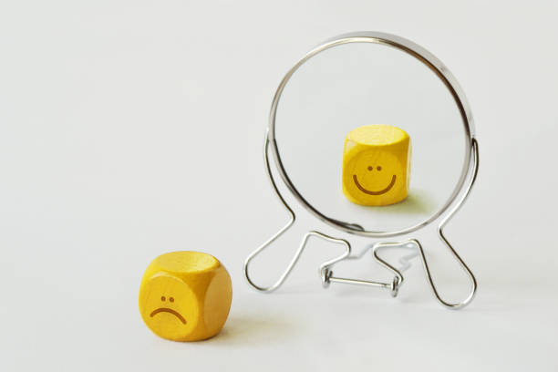 yellow dice with sad expression looking in the mirror and seeing himself smiling - concept of person who hides his pain behind his smile - sadness depression smiley face happiness imagens e fotografias de stock