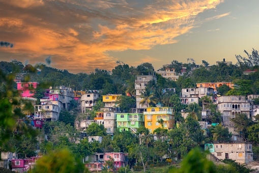 View of colorful houses on hilly area of Jamaica with lush foliage and a cloudy sky.
