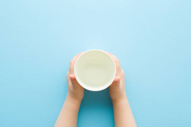 mani da bambino che tengono un bicchiere di plastica bianca di acqua dolce su sfondo di tavolo azzurro. colore pastello. primo piano. punto di vista girato. bevanda giornaliera. vista dall'alto verso il basso. - baby cup foto e immagini stock