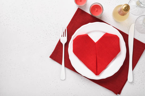 Valentinas day table setting white color with white silverware, red napkin folded as heart, candles, champagne, glasses on white background for greetings. Romantic dinner or menu concept. Mock up. stock photo