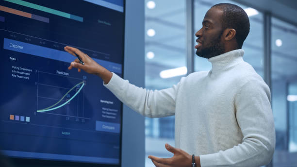 istock Diverse Modern Office: Successful Black Digital Entrepreneur Uses Tablet Computer and TV Screen with Big Data, Statistics, Talks about Company Growth. Investor, Top Managers, Executives Discuss Graphs 1363840931