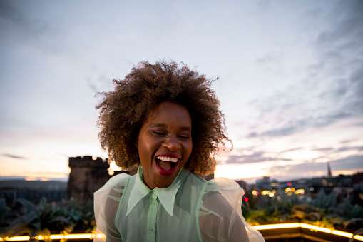 A portrait shot of a mature black woman with a large afro on a rooftop bar in Newcastle Upon Tyne. She is dancing on a table as the sun sets over the city.