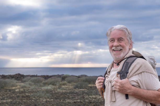 heureux grand-père aîné profitant d’une excursion en plein air en mer tenant un sac à dos. horizon au-dessus de l’eau et ciel spectaculaire avec rayon de soleil - senior adult outdoors wellbeing sky photos et images de collection