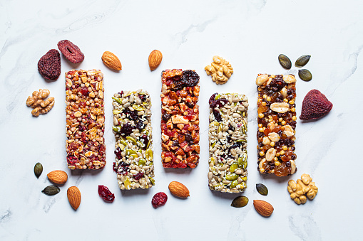 Energy granola bars with different seeds, nuts and dried fruits and berries on a white marble background, top view, copy space. Healthy snack concept.