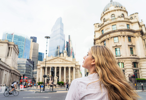 A woman in front of a junction at the heart of the City of London, with the Bank of England at left, and office buildings and skyscrapers on the horizon.
