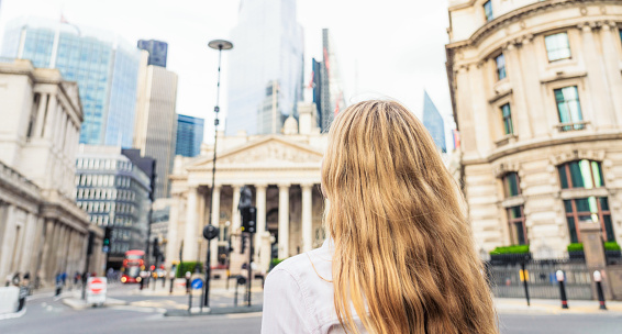 Rear view of a young woman looking up towards the towers in the heart of the City of London, the UK's key financial district, with the Bank of England at left.