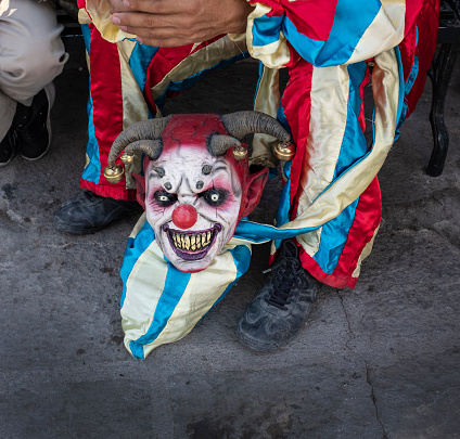 The ghoulish harlequin style mask of a Los Locos reveller laying on the ground by his feet as he sits taking a break -at the annual \