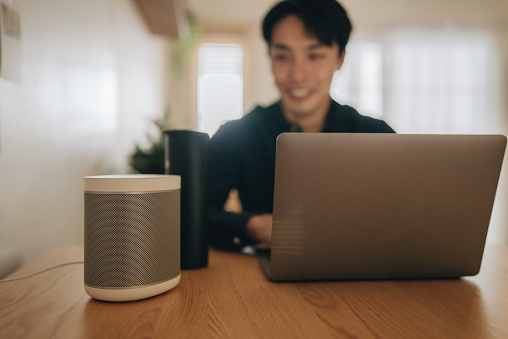 Young Asian Man Sitting at His Desk Works on Laptop, Beside Him Smart Speaker with AI Assistance Answers Questions, Plays Music, Podcast.
