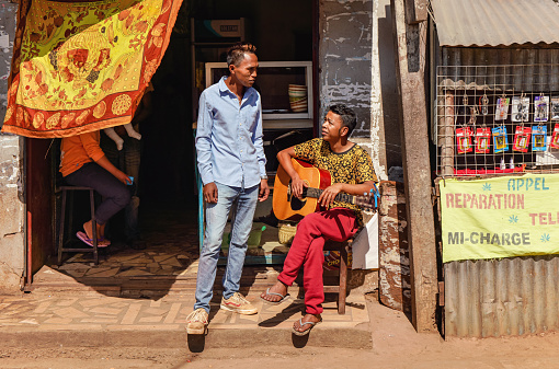 Antananarivo, Madagascar - May 07, 2019: Two unknown Malagasy men chatting in front of small shop at main street, sitting one holding guitar