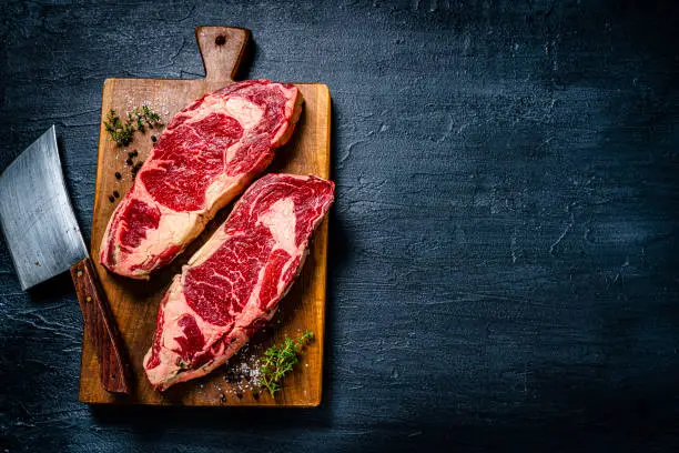 Food backgrounds: overhead view of two raw juicy beef steak fillets on a cutting board shot on black table. The composition is at the left of an horizontal frame leaving useful copy space for text and/or logo at the right. High resolution 42Mp studio digital capture taken with SONY A7rII and Zeiss Batis 40mm F2.0 CF lens