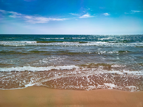 Strip of sandy beach overlooking the foaming waves of the ocean and the blue sky with clouds over the horizon - coastline and sand beach. Sunny beach and sea with clear sky. Photo of sea waves. Foaming ocean waves. Clear water. The seabed. The seashore. Sea foam. Waves on the river. Water in the lake.