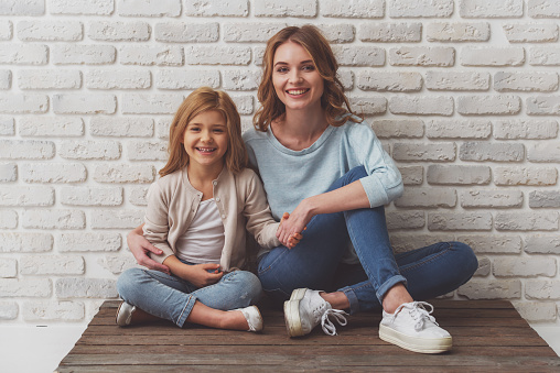 Pretty little girl and her beautiful young mother, looking in camera and smiling while sitting against white brick wall