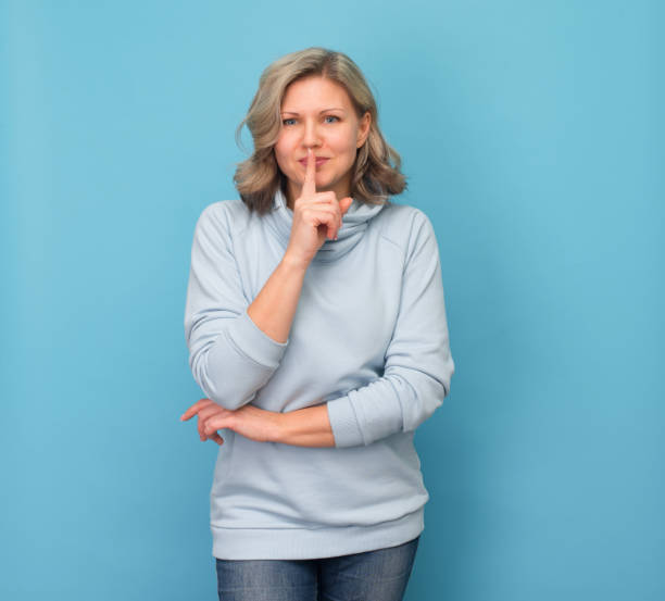 mujer risueña haciendo un gesto de silencio. atractiva mujer rubia que se lleva el dedo a los labios y pide silencio sobre fondo azul. - finger on lips whispering secrecy women fotografías e imágenes de stock