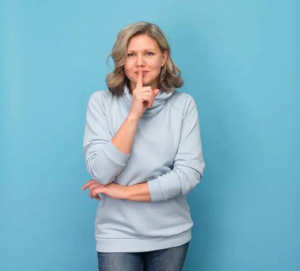 Photo of Laughing woman doing a silence gesture. Attractive blond woman putting finger up to lips and ask silence on blue background.