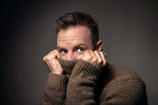 Close-up of a young man pulling a sweater over his face. Man covering face with a turtleneck sweater and looking away against grat background.