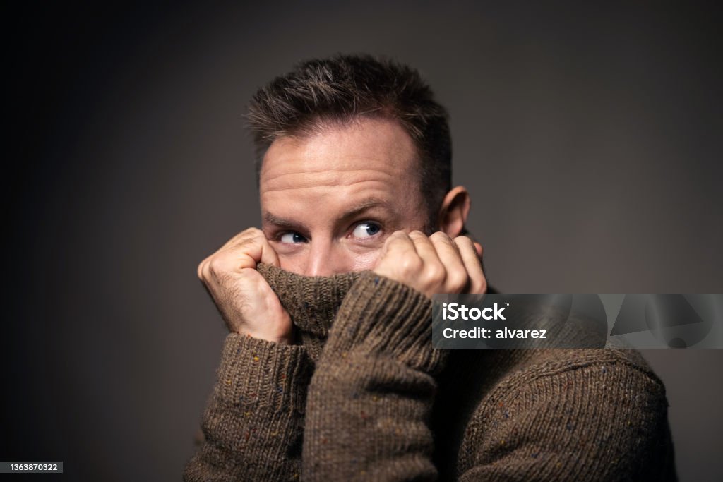 Young man covering face with turtleneck sweater Close-up of a young man pulling a sweater over his face. Man covering face with a turtleneck sweater and looking away against grat background. Embarrassment Stock Photo