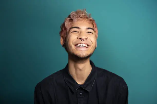 Cheerful young man smiling on blue background. Portrait of an asian man with pink hair and piercing in the studio.