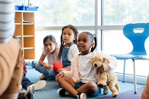 A group of attentive kindergarten students smile as they listen to their unrecognizable teacher read a book.