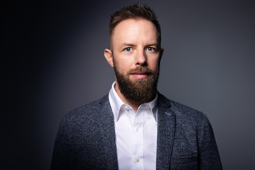 Desaturated serious bearded hipster looking away. High contrast low key dark shadow portrait isolated over black background.