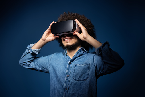 Young man using vr glasses on blue background. Middle eastern man wearing virtual reality simulator headset on studio background.