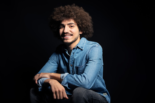 Smiling portrait of a young man looking at camera during a portrait session. Middle eastern man in casuals and curly hair sitting against black background.