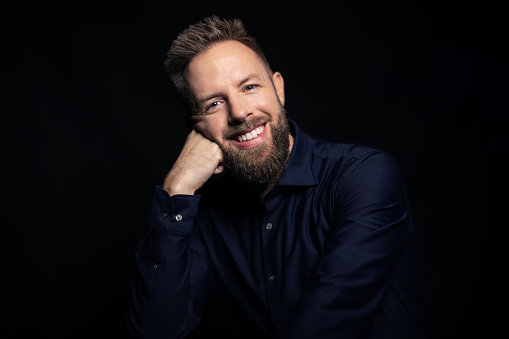 Portrait of a happy young man with beard on black background. Handsome young man with hand hand on chin and smiling at the camera.