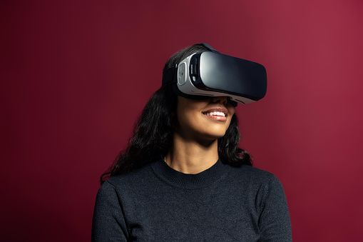 Young woman using vr glasses on red background. Latin american woman wearing virtual reality simulator headset on studio background.