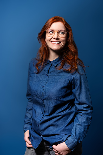 Portrait of a cheerful mature woman with red hair looking away and smiling on a blue background. Photo session of a female wearing denim shirt and hands in pocket in the studio.