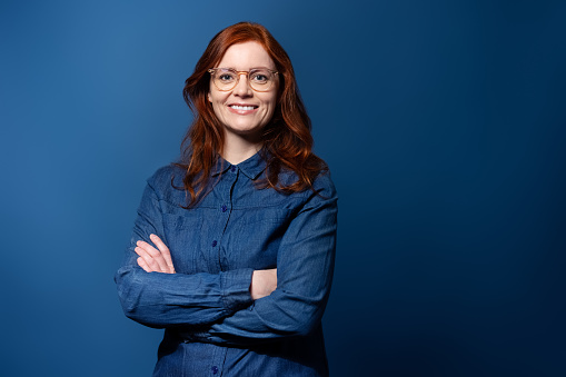 Portrait of happy mature woman with arms crossed looking at camera confidently. Woman in denim shirt standing against blue studio background and smiling.