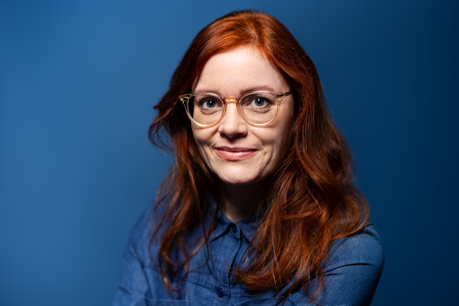 Studio portrait of confident mature woman with red hair wearing eyeglasses. Female in denim shirt standing looking at camera on blue background.